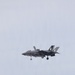 A single-seat military jet fighter is photographed in flight against an overcast gray sky. The aircraft has its landing gear deployed, suggesting it is in an approach or low-speed flight configuration. Markings and tail art indicate it is a U.S. Marine Corps aircraft.