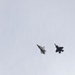 Two fifth-generation stealth fighter jets are flying in close formation against an overcast, pale grey sky. Both aircraft display the distinctive delta-wing and blended fuselage profile consistent with the F-35 Lightning II. The aircraft appear to be photographed from below at a slight angle, showing their ventral surfaces.