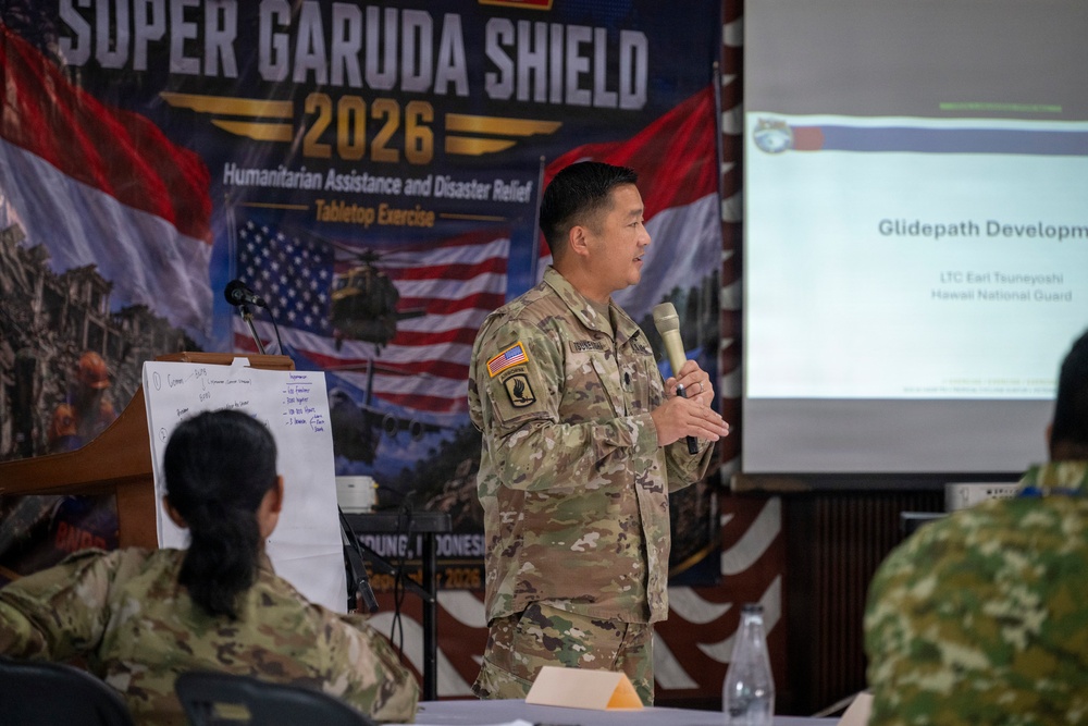 A U.S. Army officer, identified on the presentation slide as LTC Earl Tsuneyoshi of the Hawaii National Guard, is presenting at the Super Garuda Shield 2026 Humanitarian Assistance and Disaster Relief Tabletop Exercise. The event appears to be held in Bandung, Indonesia, based on partial text visible on the banner. Multiple military personnel from different nations are seated as attendees.
