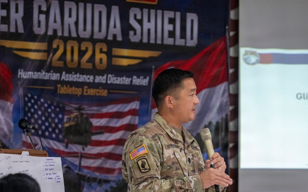 A U.S. Army officer, identified on the presentation slide as LTC Earl Tsuneyoshi of the Hawaii National Guard, is presenting at the Super Garuda Shield 2026 Humanitarian Assistance and Disaster Relief Tabletop Exercise. The event appears to be held in Bandung, Indonesia, based on partial text visible on the banner. Multiple military personnel from different nations are seated as attendees.