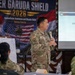 A U.S. Army officer, identified on the presentation slide as LTC Earl Tsuneyoshi of the Hawaii National Guard, is presenting at the Super Garuda Shield 2026 Humanitarian Assistance and Disaster Relief Tabletop Exercise. The event appears to be held in Bandung, Indonesia, based on partial text visible on the banner. Multiple military personnel from different nations are seated as attendees.