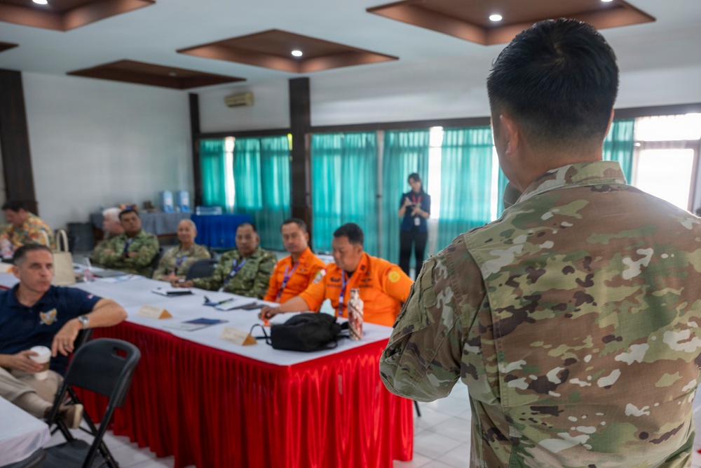 A military briefing or meeting is taking place in a conference room, with a soldier in multicam/OCP camouflage uniform addressing a seated audience. The attendees include personnel in various uniforms including orange emergency/disaster response vests, military camouflage, and civilian attire. The setting appears to be a multinational or joint coordination meeting, likely related to disaster response or humanitarian assistance.