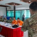 A military briefing or meeting is taking place in a conference room, with a soldier in multicam/OCP camouflage uniform addressing a seated audience. The attendees include personnel in various uniforms including orange emergency/disaster response vests, military camouflage, and civilian attire. The setting appears to be a multinational or joint coordination meeting, likely related to disaster response or humanitarian assistance.