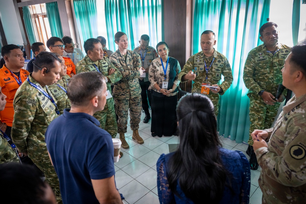 A multinational military and civilian briefing or coordination meeting is taking place indoors, likely in Southeast Asia. Personnel in various camouflage uniforms, including those marked 'TNI AD' (Indonesian Army) and what appears to be U.S. military uniforms, are gathered in a circle. Civilian participants wearing lanyards and ID badges are also present, suggesting an interagency or joint exercise environment.