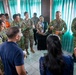 A multinational military and civilian briefing or coordination meeting is taking place indoors, likely in Southeast Asia. Personnel in various camouflage uniforms, including those marked 'TNI AD' (Indonesian Army) and what appears to be U.S. military uniforms, are gathered in a circle. Civilian participants wearing lanyards and ID badges are also present, suggesting an interagency or joint exercise environment.