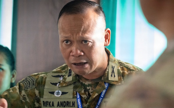 A military officer wearing a camouflage uniform with the name tape 'RIFKA ANDRI.H' is engaged in an animated discussion, holding papers and a pen. He is wearing rank insignia and multiple unit patches, along with a blue lanyard with an ID badge. The setting appears to be an indoor military briefing or coordination meeting with other personnel present.