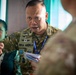 A military officer wearing a camouflage uniform with the name tape 'RIFKA ANDRI.H' is engaged in an animated discussion, holding papers and a pen. He is wearing rank insignia and multiple unit patches, along with a blue lanyard with an ID badge. The setting appears to be an indoor military briefing or coordination meeting with other personnel present.