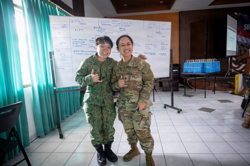 Two female military personnel in different camouflage uniforms pose together smiling in what appears to be a conference or briefing room. One wears a Singapore Armed Forces (SAF) uniform with a 'SINGAPORE' patch, while the other wears a US Army OCP (Operational Camouflage Pattern) uniform. Both are making shaka/thumbs-up hand gestures, suggesting a friendly bilateral military engagement or exercise.