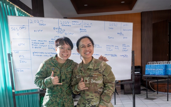Two female military personnel in different camouflage uniforms pose together smiling in what appears to be a conference or briefing room. One wears a Singapore Armed Forces (SAF) uniform with a 'SINGAPORE' patch, while the other wears a US Army OCP (Operational Camouflage Pattern) uniform. Both are making shaka/thumbs-up hand gestures, suggesting a friendly bilateral military engagement or exercise.