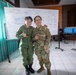 Two female military personnel in different camouflage uniforms pose together smiling in what appears to be a conference or briefing room. One wears a Singapore Armed Forces (SAF) uniform with a 'SINGAPORE' patch, while the other wears a US Army OCP (Operational Camouflage Pattern) uniform. Both are making shaka/thumbs-up hand gestures, suggesting a friendly bilateral military engagement or exercise.