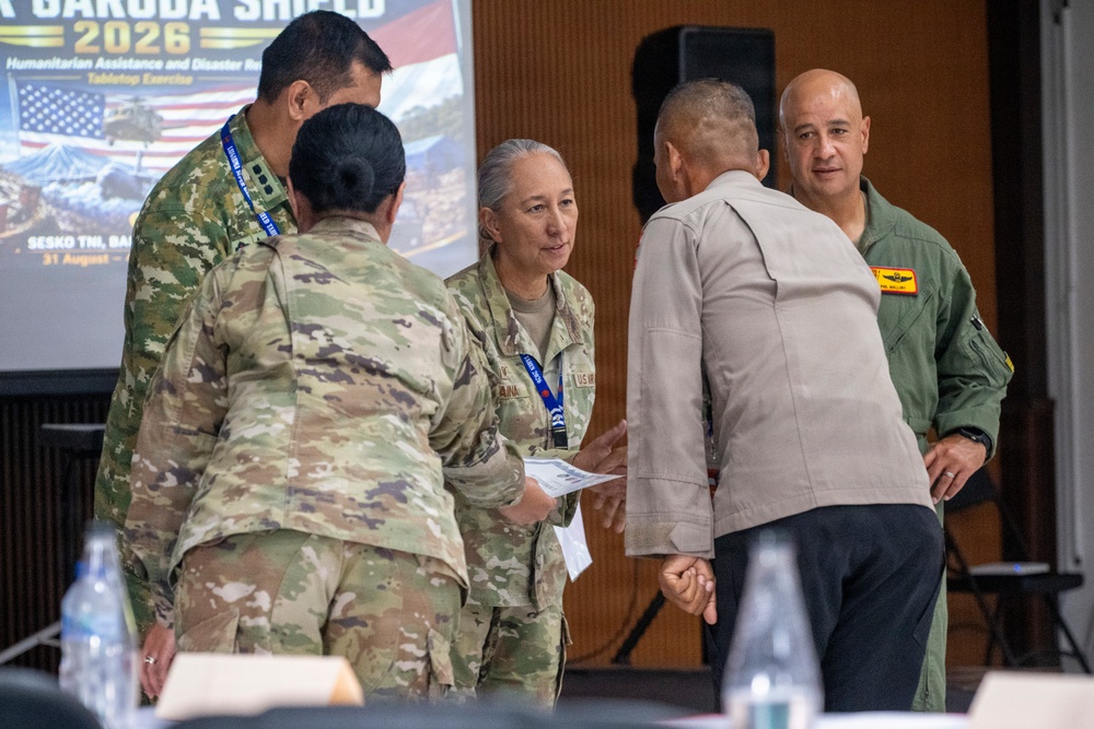 Military personnel from multiple nations are gathered at the Garuda Shield 2026 Humanitarian Assistance and Disaster Relief Tabletop Exercise, held at SESKO TNI, Bandung, starting August 31. Several uniformed individuals are engaged in conversation and what appears to be a document or certificate exchange. The event involves both U.S. military personnel in OCP camouflage and individuals in other military uniforms, suggesting a multinational joint exercise.