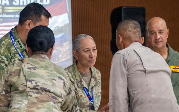 Military personnel from multiple nations are gathered at the Garuda Shield 2026 Humanitarian Assistance and Disaster Relief Tabletop Exercise, held at SESKO TNI, Bandung, starting August 31. Several uniformed individuals are engaged in conversation and what appears to be a document or certificate exchange. The event involves both U.S. military personnel in OCP camouflage and individuals in other military uniforms, suggesting a multinational joint exercise.