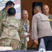 Military personnel from multiple nations are gathered at the Garuda Shield 2026 Humanitarian Assistance and Disaster Relief Tabletop Exercise, held at SESKO TNI, Bandung, starting August 31. Several uniformed individuals are engaged in conversation and what appears to be a document or certificate exchange. The event involves both U.S. military personnel in OCP camouflage and individuals in other military uniforms, suggesting a multinational joint exercise.