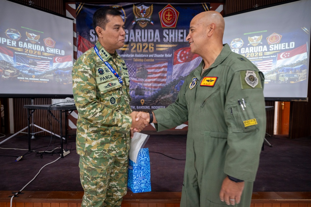 Two military officers are shaking hands at the Super Garuda Shield 2026 exercise event held at Sesko TNI, Bandung, Indonesia. The event focuses on Humanitarian Assistance and Disaster Relief (HADR) as a Tabletop Exercise. One officer wears Indonesian TNI camouflage uniform with a name tag reading 'PANCA', while the other wears an olive green flight suit.
