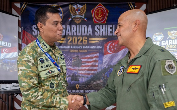 Two military officers are shaking hands at the Super Garuda Shield 2026 exercise event held at Sesko TNI, Bandung, Indonesia. The event focuses on Humanitarian Assistance and Disaster Relief (HADR) as a Tabletop Exercise. One officer wears Indonesian TNI camouflage uniform with a name tag reading 'PANCA', while the other wears an olive green flight suit.