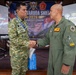 Two military officers are shaking hands at the Super Garuda Shield 2026 exercise event held at Sesko TNI, Bandung, Indonesia. The event focuses on Humanitarian Assistance and Disaster Relief (HADR) as a Tabletop Exercise. One officer wears Indonesian TNI camouflage uniform with a name tag reading 'PANCA', while the other wears an olive green flight suit.