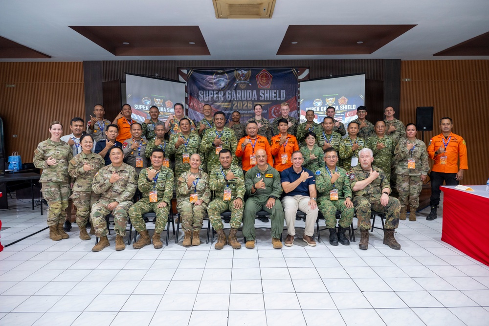 A large group of multinational military and civilian personnel pose for a formal group photograph at the 'Super Garuda Shield 2026' exercise or event. Participants are wearing various uniforms including US Army multicam, Indonesian military camouflage, orange search-and-rescue suits, and civilian attire. The group is arranged in three rows inside an indoor venue with a branded backdrop.