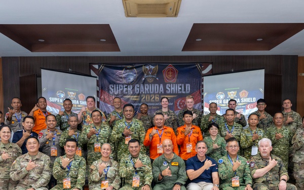 A large group of multinational military and civilian personnel pose for a formal group photograph at the 'Super Garuda Shield 2026' exercise or event. Participants are wearing various uniforms including US Army multicam, Indonesian military camouflage, orange search-and-rescue suits, and civilian attire. The group is arranged in three rows inside an indoor venue with a branded backdrop.