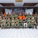 A large group of multinational military and civilian personnel pose for a formal group photograph at the 'Super Garuda Shield 2026' exercise or event. Participants are wearing various uniforms including US Army multicam, Indonesian military camouflage, orange search-and-rescue suits, and civilian attire. The group is arranged in three rows inside an indoor venue with a branded backdrop.