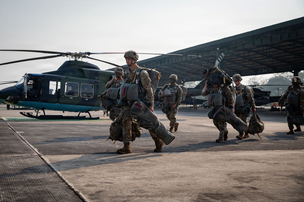 A group of military personnel in full combat gear and parachute equipment are walking across a tarmac or airfield apron. They are carrying large parachute packs and equipment bags, moving away from or toward a dark green Bell 412 or similar utility helicopter parked nearby. A large aircraft hangar structure is visible in the background.
