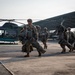 A group of military personnel in full combat gear and parachute equipment are walking across a tarmac or airfield apron. They are carrying large parachute packs and equipment bags, moving away from or toward a dark green Bell 412 or similar utility helicopter parked nearby. A large aircraft hangar structure is visible in the background.