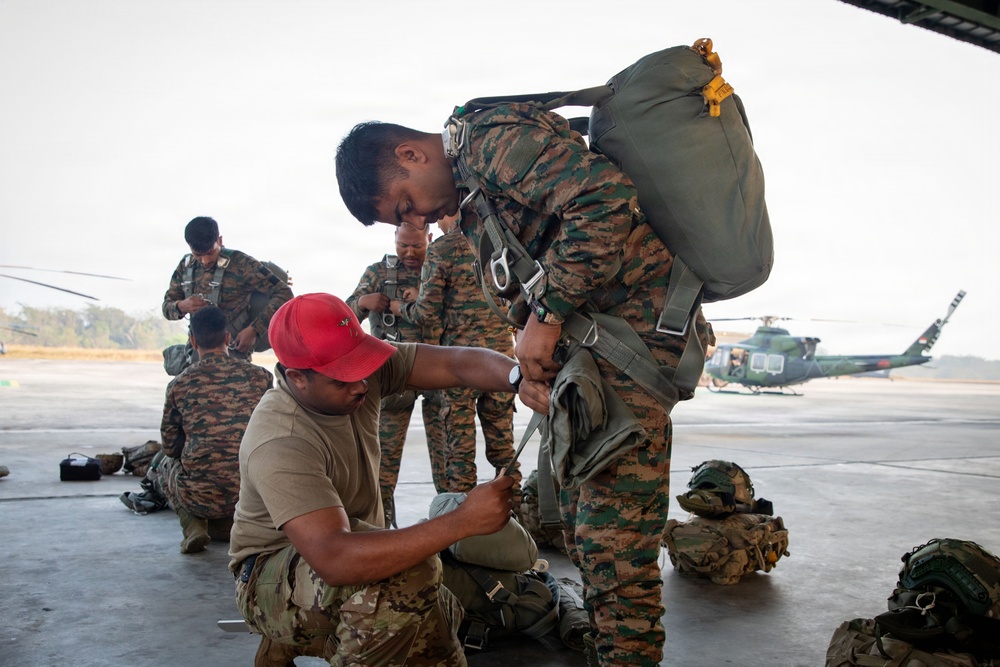Military personnel are preparing parachute or rappelling equipment on a tarmac or hangar apron. A kneeling soldier in a red cap and tan uniform is assisting a standing soldier in camouflage with harness adjustments. A helicopter, likely a UH-1 Huey variant, is visible in the background along with additional soldiers conducting similar equipment checks.