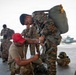Military personnel are preparing parachute or rappelling equipment on a tarmac or hangar apron. A kneeling soldier in a red cap and tan uniform is assisting a standing soldier in camouflage with harness adjustments. A helicopter, likely a UH-1 Huey variant, is visible in the background along with additional soldiers conducting similar equipment checks.
