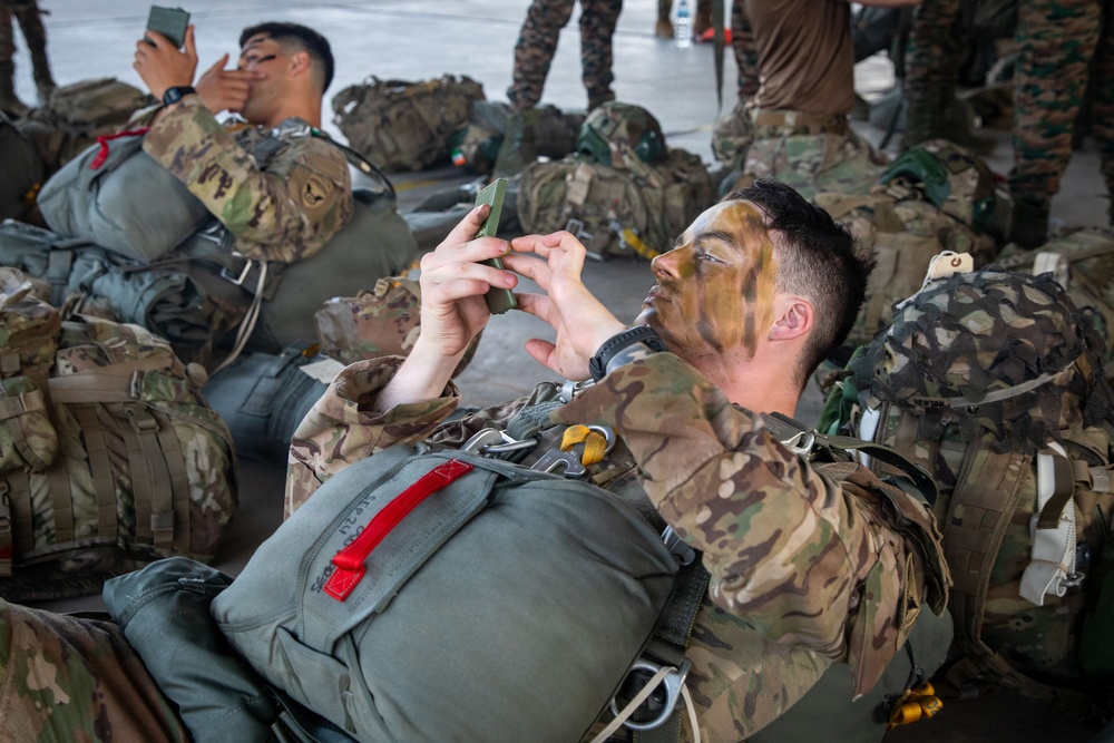 Military personnel in full combat gear are resting on what appears to be a hangar or staging area floor, surrounded by packed rucksacks and parachute equipment. The foreground soldier has camouflage face paint applied and is using a mobile phone, while wearing a parachute harness with visible red deployment handle. Multiple soldiers in various camouflage uniforms are visible in the background, suggesting a pre-jump or pre-mission staging operation.