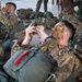 Military personnel in full combat gear are resting on what appears to be a hangar or staging area floor, surrounded by packed rucksacks and parachute equipment. The foreground soldier has camouflage face paint applied and is using a mobile phone, while wearing a parachute harness with visible red deployment handle. Multiple soldiers in various camouflage uniforms are visible in the background, suggesting a pre-jump or pre-mission staging operation.