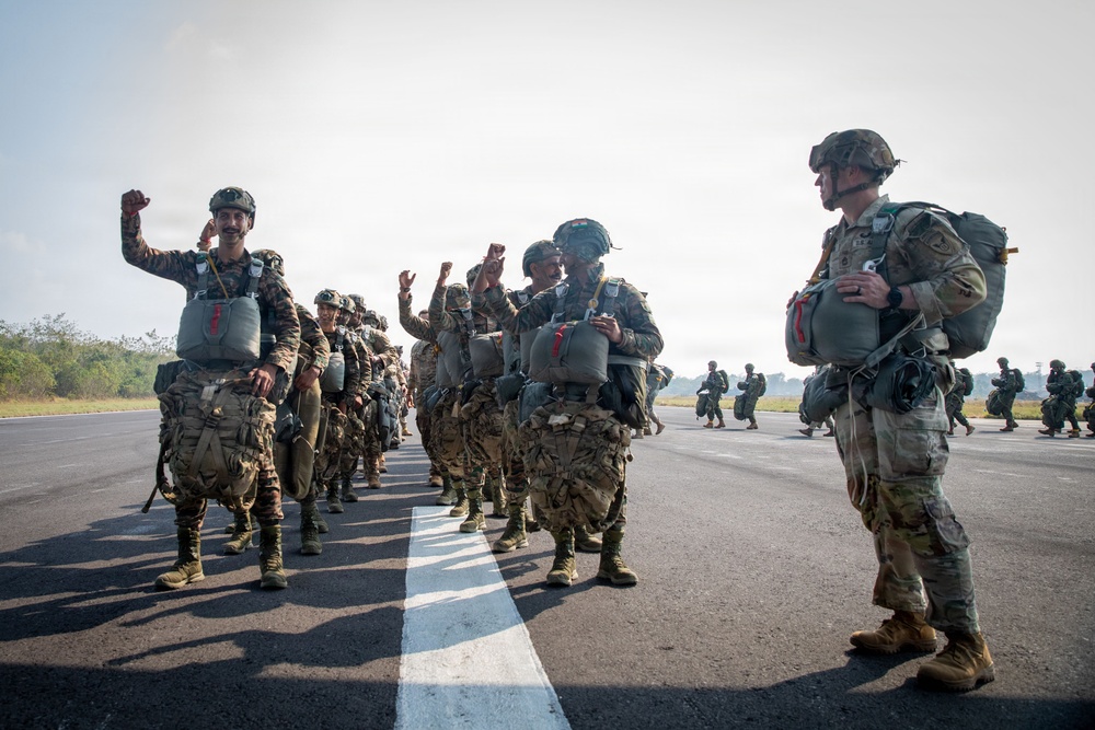 A group of military paratroopers in full jump gear are assembled on an airfield tarmac, appearing to be in high spirits before or after a parachute operation. The soldiers are wearing mixed camouflage patterns suggesting a multinational or joint military exercise. Several soldiers are raising their fists in celebration or motivation while lined up along a runway centerline.