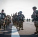 A group of military paratroopers in full jump gear are assembled on an airfield tarmac, appearing to be in high spirits before or after a parachute operation. The soldiers are wearing mixed camouflage patterns suggesting a multinational or joint military exercise. Several soldiers are raising their fists in celebration or motivation while lined up along a runway centerline.