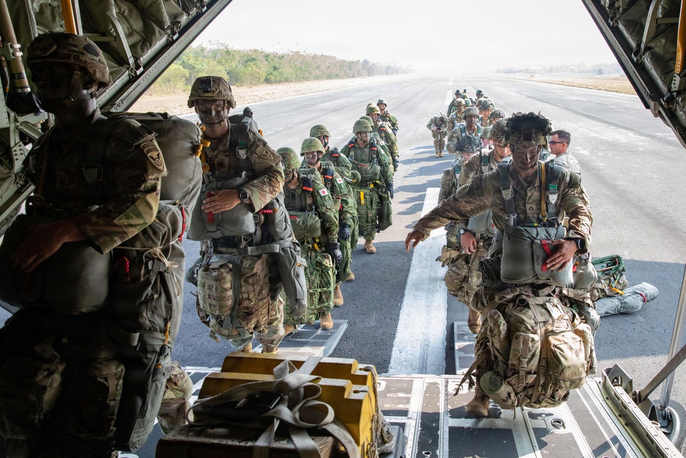 Military personnel in full parachute gear are boarding a large military transport aircraft, likely a C-130 Hercules, via its rear cargo ramp on an airstrip. The group includes soldiers wearing at least two distinct camouflage patterns, suggesting a multinational or joint military exercise. The scene depicts a pre-jump load operation with troops carrying parachute packs and tactical equipment.