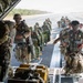 Military personnel in full parachute gear are boarding a large military transport aircraft, likely a C-130 Hercules, via its rear cargo ramp on an airstrip. The group includes soldiers wearing at least two distinct camouflage patterns, suggesting a multinational or joint military exercise. The scene depicts a pre-jump load operation with troops carrying parachute packs and tactical equipment.