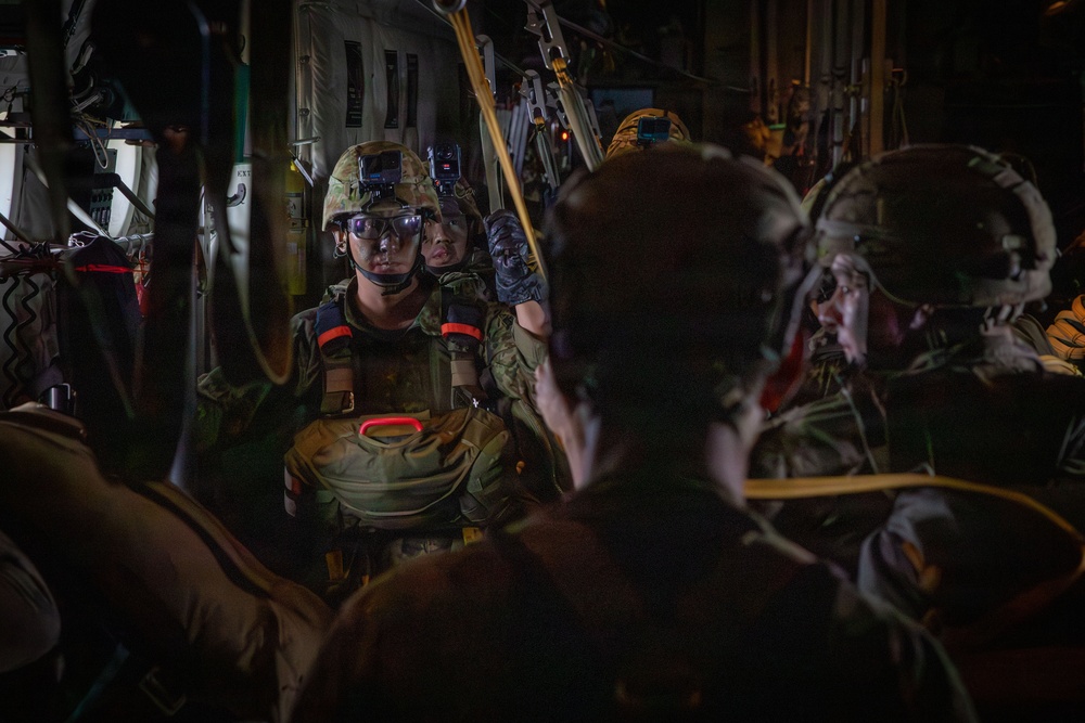 Military personnel are gathered inside what appears to be a military transport aircraft, likely preparing for a parachute jump operation. The soldiers are equipped with full tactical gear including helmets, protective eyewear, body armor, and parachute harnesses. The interior lighting is dim, suggesting either nighttime operations or low-light conditions inside the aircraft.