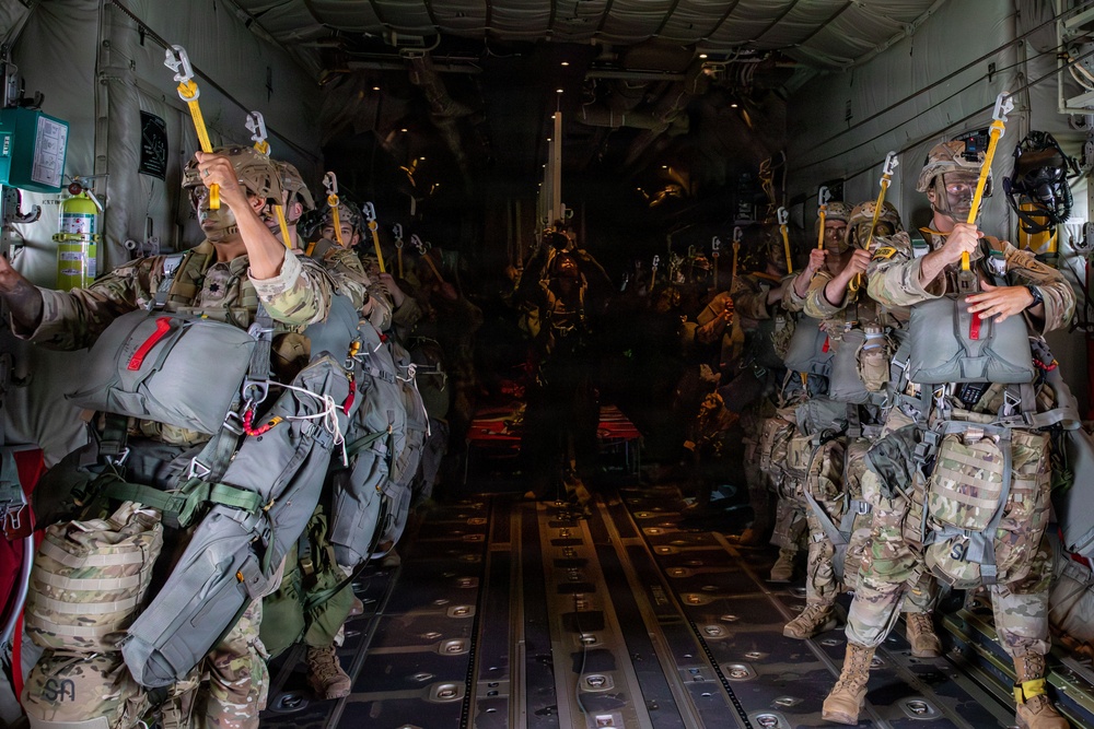 Military paratroopers are lined up in two rows inside a large military transport aircraft, preparing for a static-line parachute jump. The soldiers are fully equipped with parachute rigs, combat gear, and helmets, and are holding yellow static-line snap hooks above their heads in preparation to attach to the anchor line cable. The interior of the aircraft is dimly lit toward the rear, with two columns of jumpers facing the center aisle.