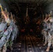 Military paratroopers are lined up in two rows inside a large military transport aircraft, preparing for a static-line parachute jump. The soldiers are fully equipped with parachute rigs, combat gear, and helmets, and are holding yellow static-line snap hooks above their heads in preparation to attach to the anchor line cable. The interior of the aircraft is dimly lit toward the rear, with two columns of jumpers facing the center aisle.