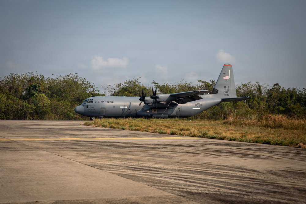 A U.S. Air Force C-130J Super Hercules military transport aircraft is parked or taxiing on a concrete runway or apron. The aircraft bears tail code 'YJ' and markings indicating it belongs to 5th Air Force, with serial number 1843 visible on the tail. The setting appears to be a tropical or subtropical airfield surrounded by dense vegetation.