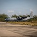 A U.S. Air Force C-130J Super Hercules military transport aircraft is parked or taxiing on a concrete runway or apron. The aircraft bears tail code 'YJ' and markings indicating it belongs to 5th Air Force, with serial number 1843 visible on the tail. The setting appears to be a tropical or subtropical airfield surrounded by dense vegetation.