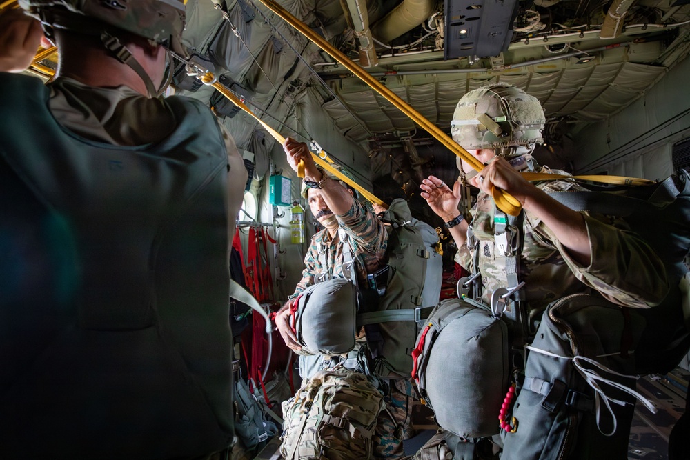 Military personnel are inside what appears to be a C-130 Hercules transport aircraft, preparing for a parachute jump operation. Soldiers wearing combat gear, helmets, and parachute rigs are attaching static lines to the anchor line cable running along the aircraft's ceiling. The scene depicts a standard airborne operation pre-jump sequence.