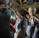 Military personnel are inside what appears to be a C-130 Hercules transport aircraft, preparing for a parachute jump operation. Soldiers wearing combat gear, helmets, and parachute rigs are attaching static lines to the anchor line cable running along the aircraft's ceiling. The scene depicts a standard airborne operation pre-jump sequence.