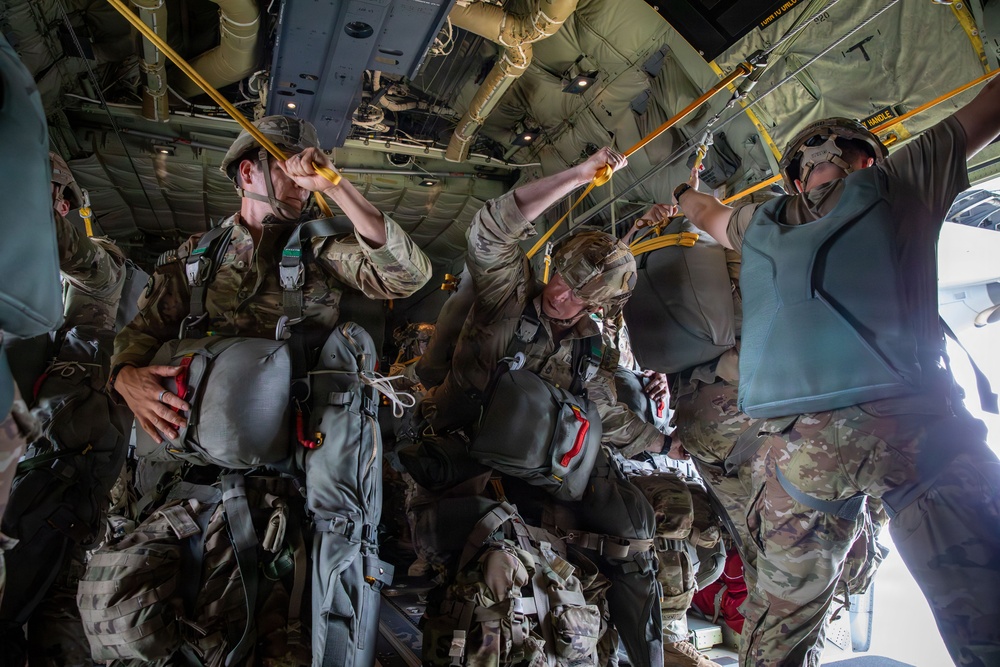 Military paratroopers are preparing for a static-line jump inside what appears to be a C-130 Hercules transport aircraft. The soldiers are fully equipped with parachute rigs and combat gear, and are hooking their static lines to the anchor line cable overhead. The aircraft's rear ramp appears to be open, indicating they are near the jump point.