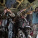 Military paratroopers are preparing for a static-line jump inside what appears to be a C-130 Hercules transport aircraft. The soldiers are fully equipped with parachute rigs and combat gear, and are hooking their static lines to the anchor line cable overhead. The aircraft's rear ramp appears to be open, indicating they are near the jump point.