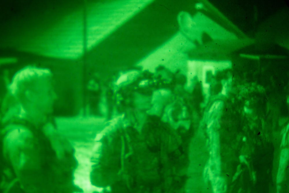 Image captured through night vision optics, showing a group of individuals in what appears to be a military or tactical nighttime operation. The characteristic green monochromatic rendering confirms the use of image intensification technology. Multiple personnel are visible near structures in low-light conditions.