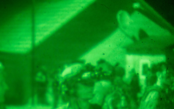 Image captured through night vision optics, showing a group of individuals in what appears to be a military or tactical nighttime operation. The characteristic green monochromatic rendering confirms the use of image intensification technology. Multiple personnel are visible near structures in low-light conditions.