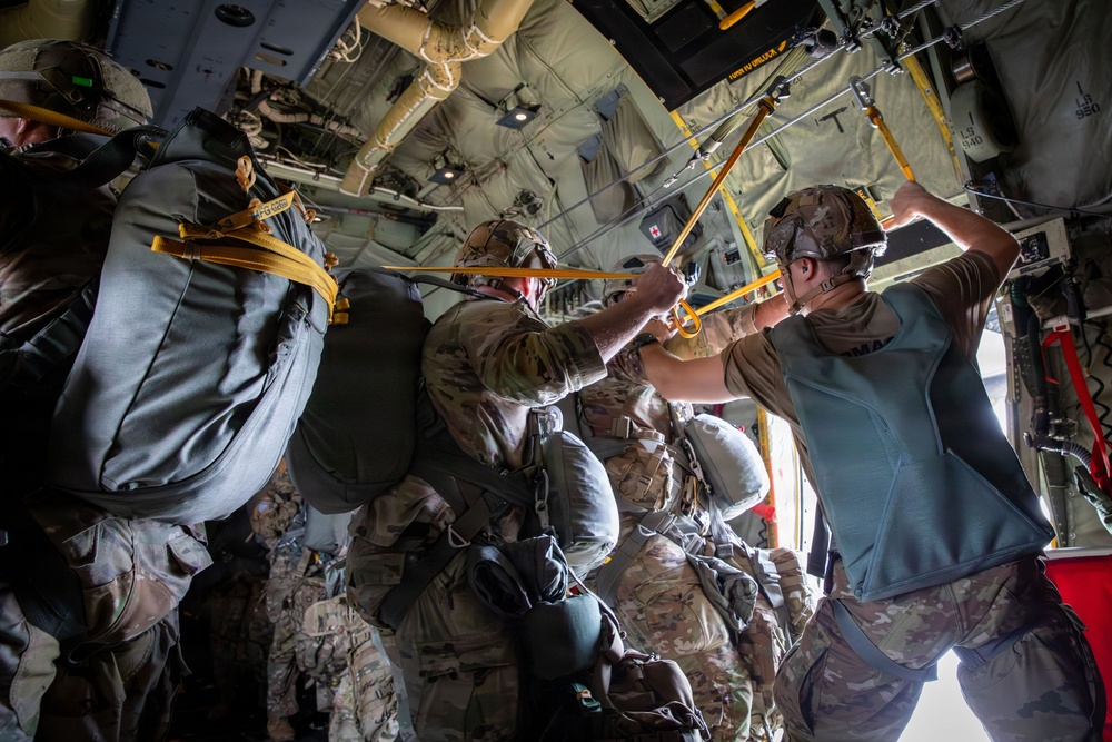 Military personnel are positioned inside what appears to be a military transport aircraft, likely a C-130 Hercules, preparing for a parachute jump operation. The soldiers are equipped with full parachute rigs and combat gear, standing near the open rear ramp of the aircraft. Yellow static lines are visible attached to an overhead anchor cable, indicating an imminent static-line parachute deployment.