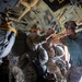Military personnel are positioned inside what appears to be a military transport aircraft, likely a C-130 Hercules, preparing for a parachute jump operation. The soldiers are equipped with full parachute rigs and combat gear, standing near the open rear ramp of the aircraft. Yellow static lines are visible attached to an overhead anchor cable, indicating an imminent static-line parachute deployment.