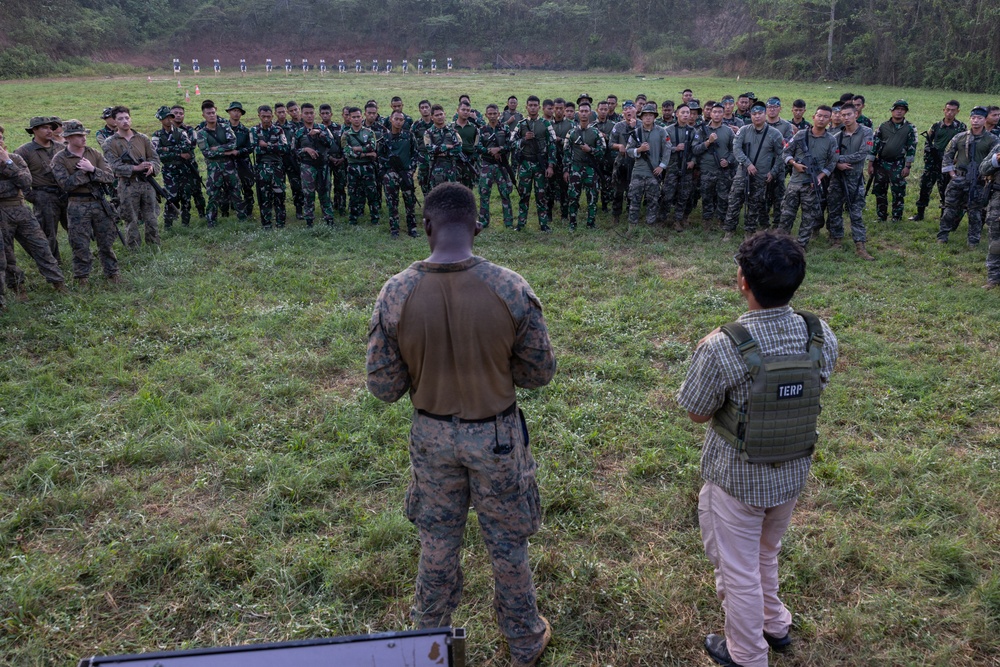 A military training or briefing event is taking place on an outdoor range or field, where a U.S. Marine in camouflage uniform addresses a large semicircular formation of soldiers in green camouflage uniforms. Beside the Marine stands an individual wearing a tactical vest labeled 'TERP' (interpreter), suggesting a multinational or joint training exercise. Shooting targets are visible in the background, indicating this is a marksmanship or weapons training range.