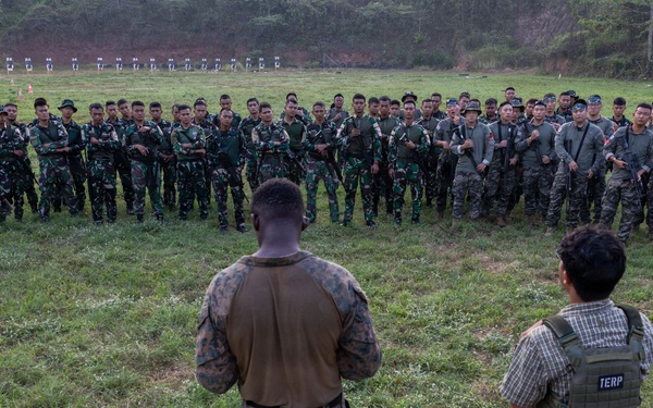 A military training or briefing event is taking place on an outdoor range or field, where a U.S. Marine in camouflage uniform addresses a large semicircular formation of soldiers in green camouflage uniforms. Beside the Marine stands an individual wearing a tactical vest labeled 'TERP' (interpreter), suggesting a multinational or joint training exercise. Shooting targets are visible in the background, indicating this is a marksmanship or weapons training range.