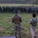 A military training or briefing event is taking place on an outdoor range or field, where a U.S. Marine in camouflage uniform addresses a large semicircular formation of soldiers in green camouflage uniforms. Beside the Marine stands an individual wearing a tactical vest labeled 'TERP' (interpreter), suggesting a multinational or joint training exercise. Shooting targets are visible in the background, indicating this is a marksmanship or weapons training range.