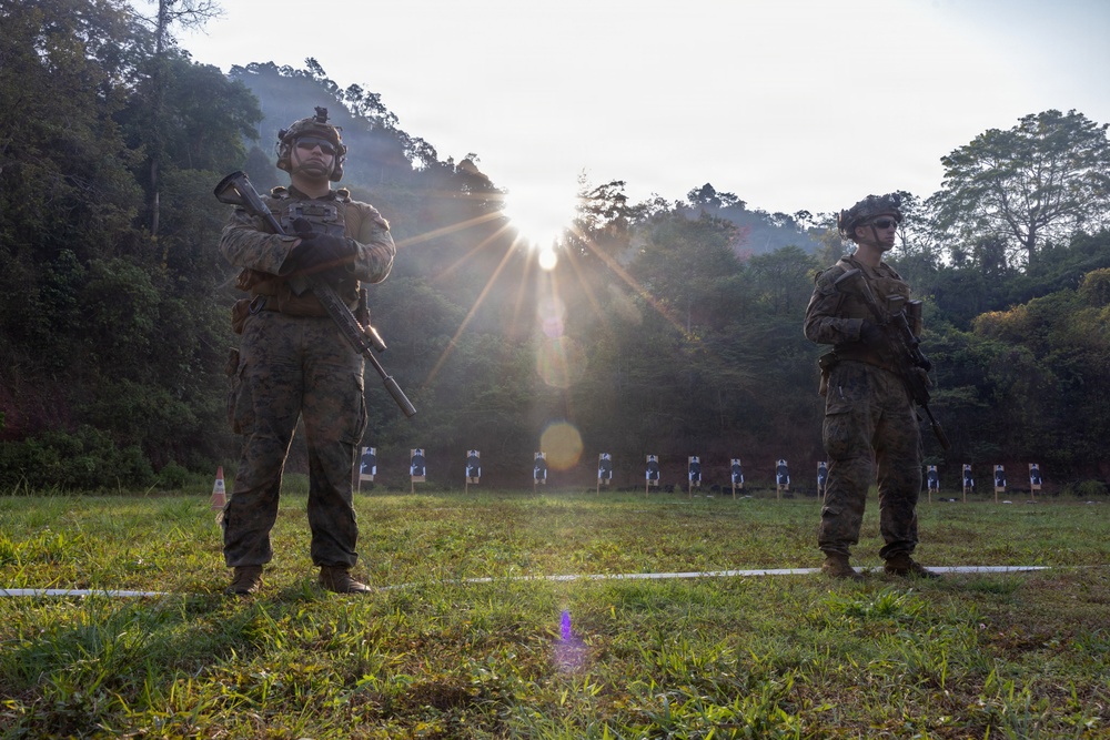 Two armed military personnel in full combat gear stand on a grass firing range, with a row of paper shooting targets visible in the background. The scene is backlit by a strong sun flare breaking through trees, creating dramatic lighting with lens flare artifacts. The setting appears to be an outdoor range in a forested, tropical or subtropical environment.